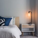Bed with white bedding and blue cushions next to a nightstand with a lit lamp
